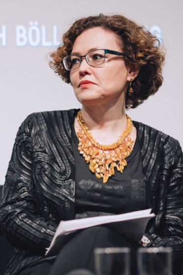 Maria Tlostanova sits in a grey suit jacket and a brown shell necklace with short curly hair and glasses. She looks to her right.