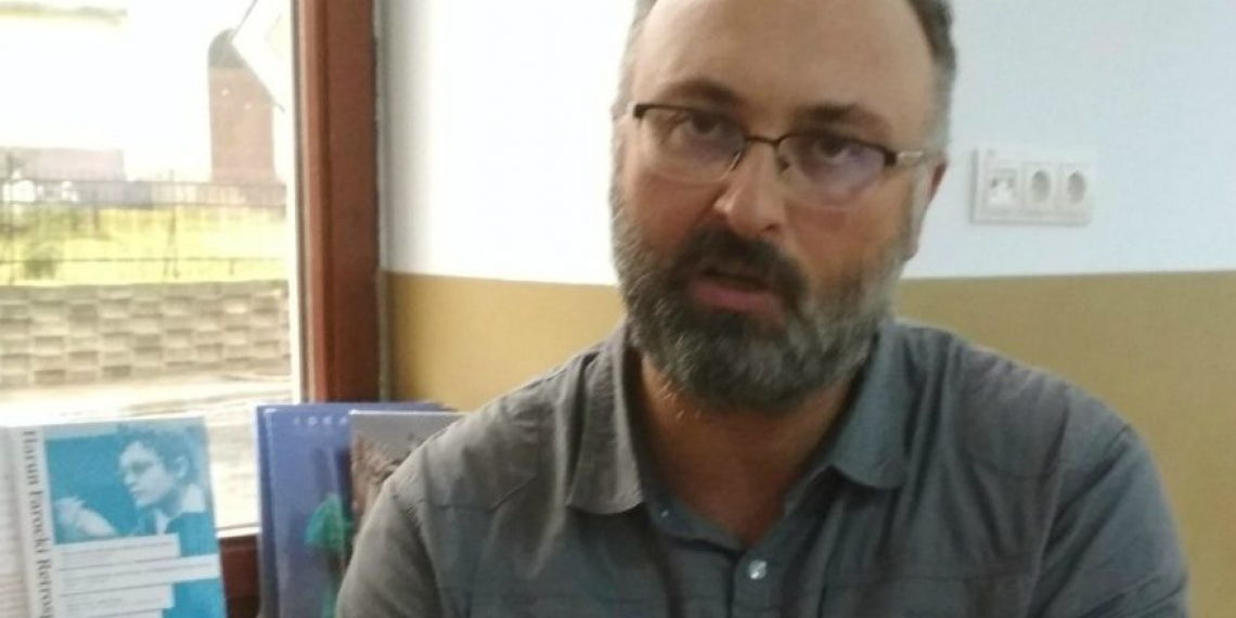 Portrait of the philosopher Ovidiu Tichindeleanu in a grey collared shirt, in the midst of talking; there are books in the background